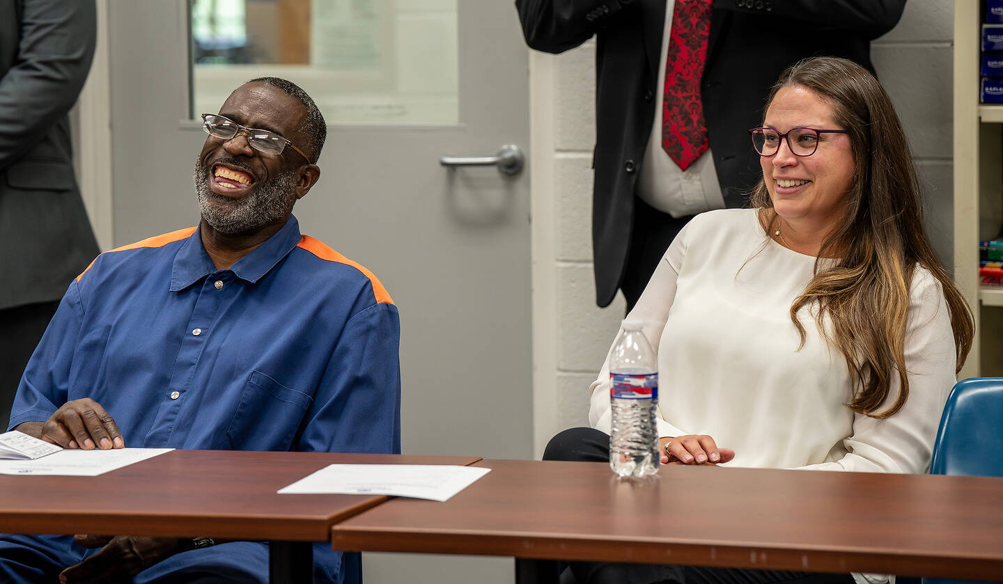 Two individuals are seated at a table, smiling and laughing during what appears to be a formal event or meeting.